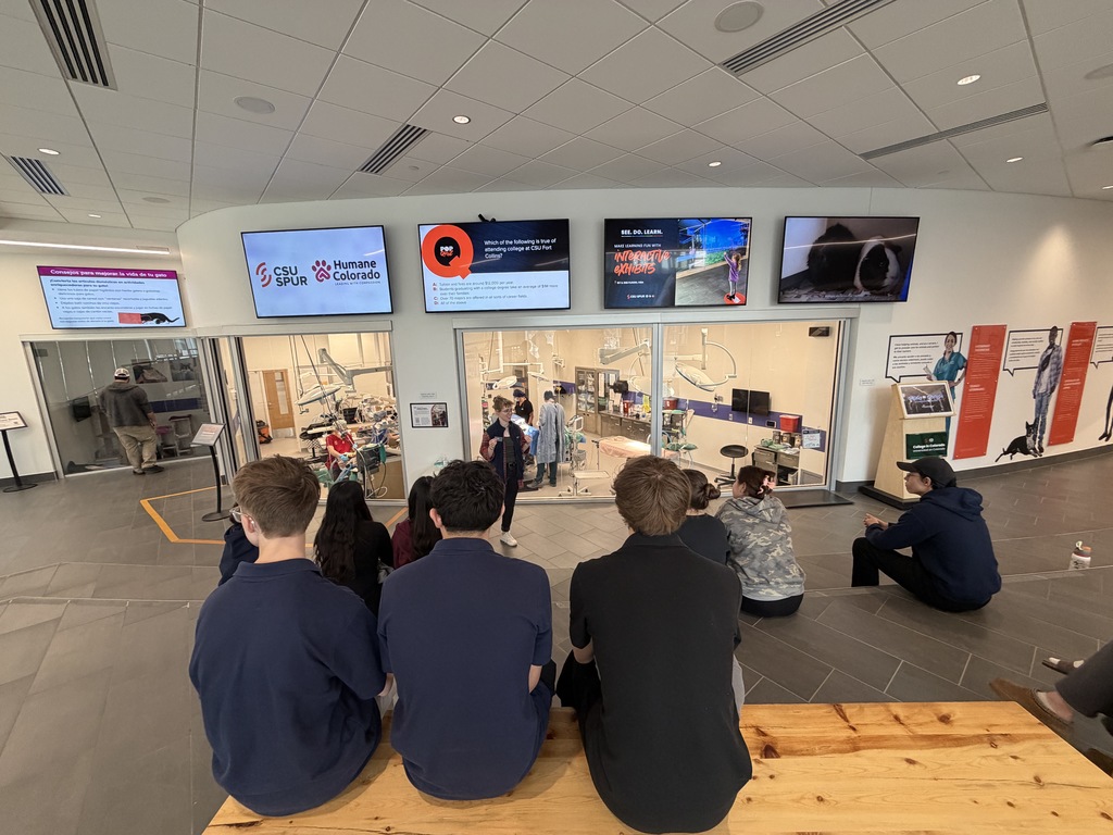 Group of high school students sits on steps facing a presenter speaking in front of large observation windows into a veterinary lab. Multiple screens above display slides, including logos for CSU Spur and Humane Colorado.