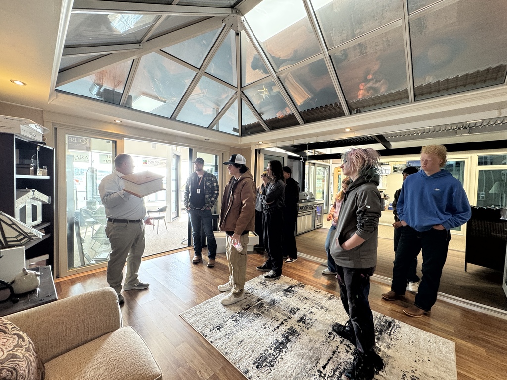 Instructor stands inside a sunroom-style display with a glass ceiling, holding a sample piece while explaining to a group of students gathered around in a home design showroom.