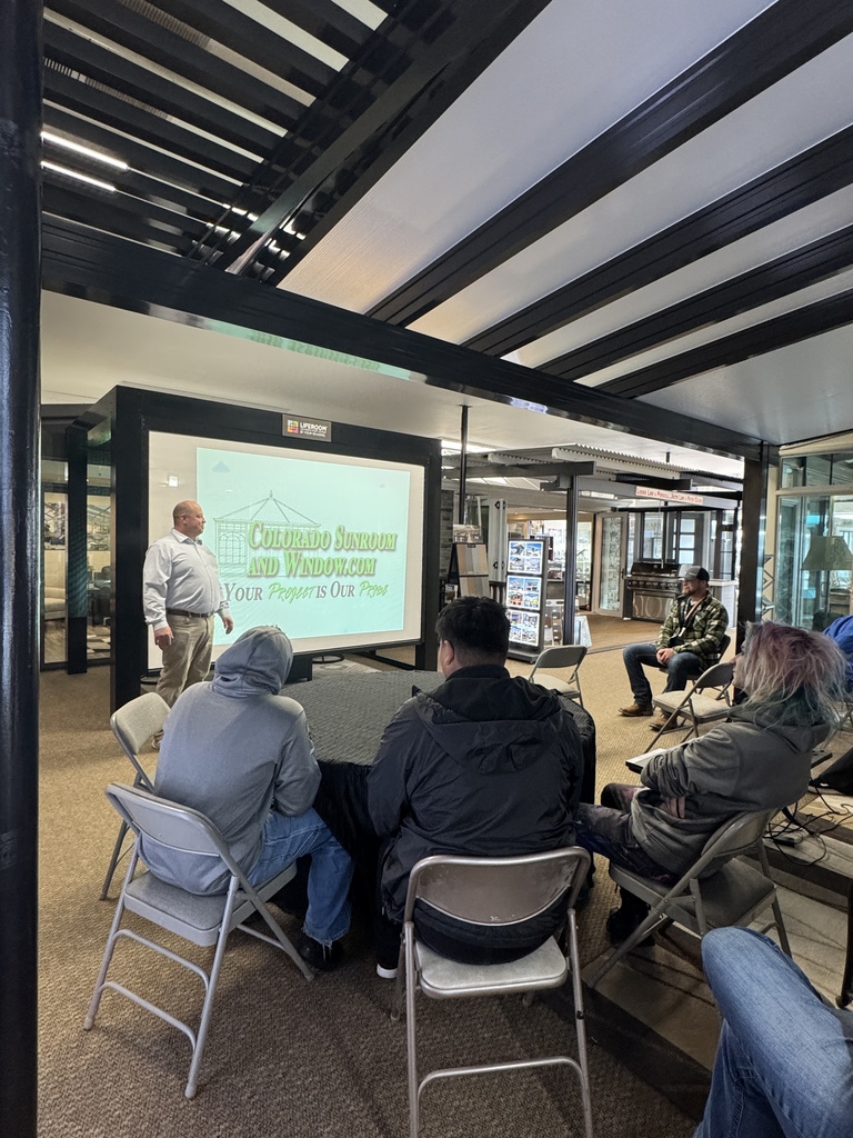 Small group of students sits on folding chairs listening to a presenter standing beside a large screen displaying “Colorado Sunroom and Window.com – Your Project is Our Pride” in a showroom setting.