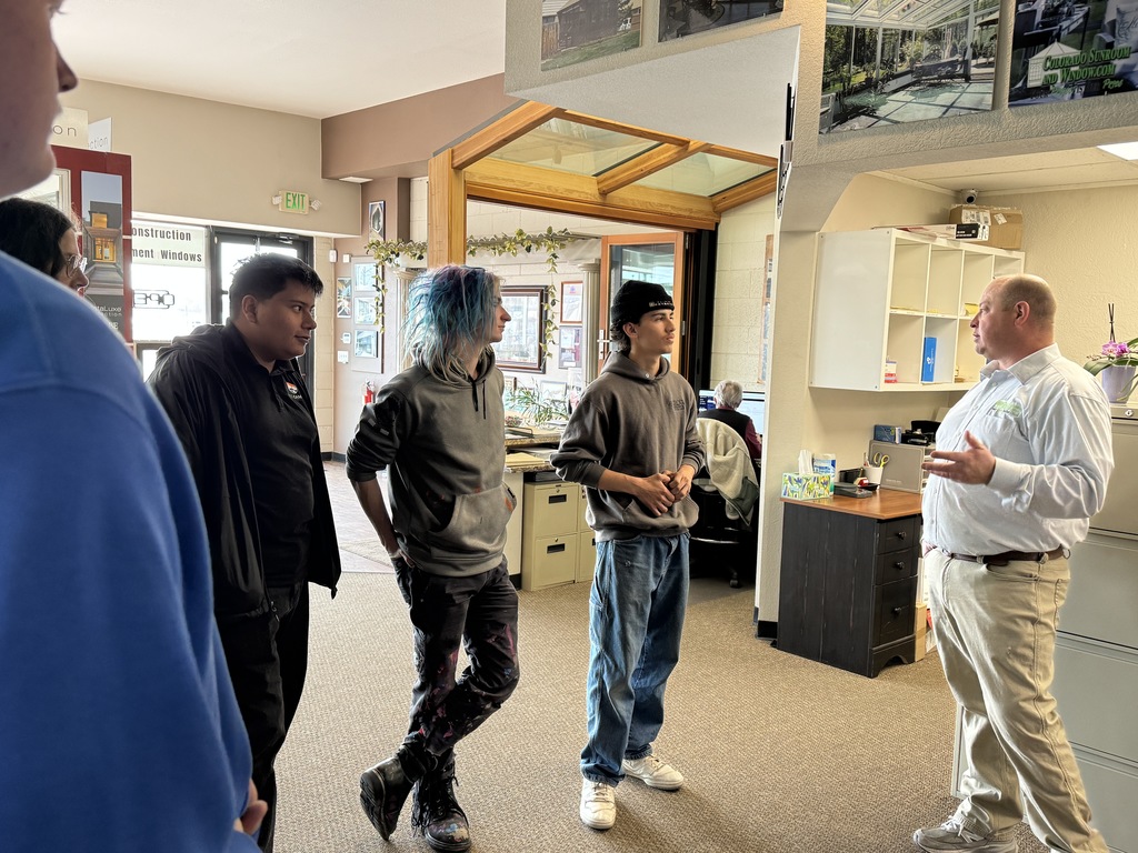 Instructor speaks with a small group of students inside a showroom office area, gesturing while the students listen attentively near displays of windows and sunroom examples.