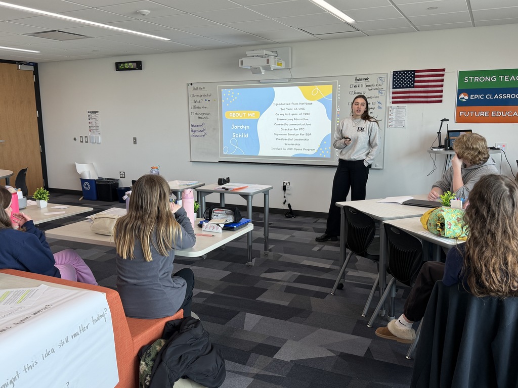 Student stands at the front of a classroom giving a presentation while classmates sit at tables and listen. A slide titled “About Me: Jordyn Schild” is projected on a screen behind them, with an American flag and classroom posters on the wall.