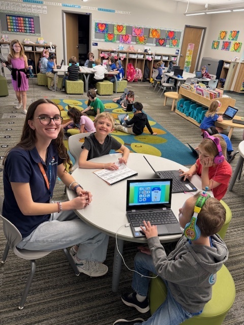 Two individuals stand in a building lobby near a staircase and a sign that reads “For privacy reasons, please wait here until called,” both smiling at the camera.