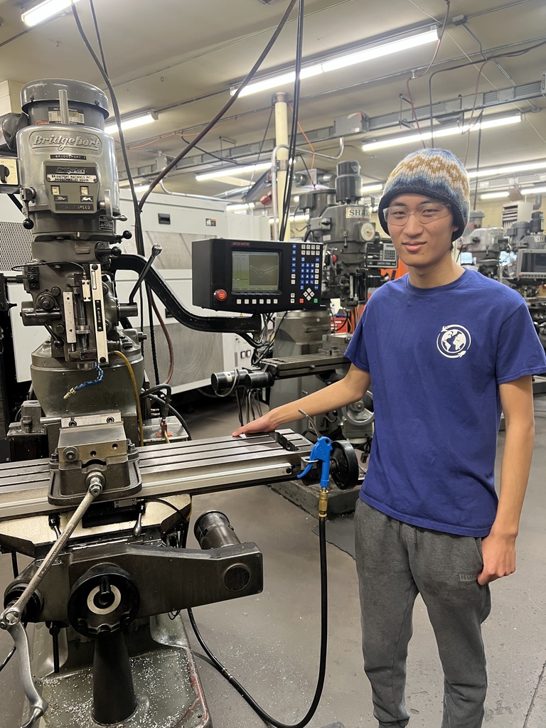 Student stands beside a large milling machine in a manufacturing lab, resting a hand on the equipment while wearing a knit hat and safety-appropriate attire.