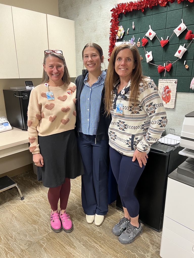 Three adult women stand together smiling in an office or classroom space decorated with red heart-themed decorations and a bulletin board behind them.