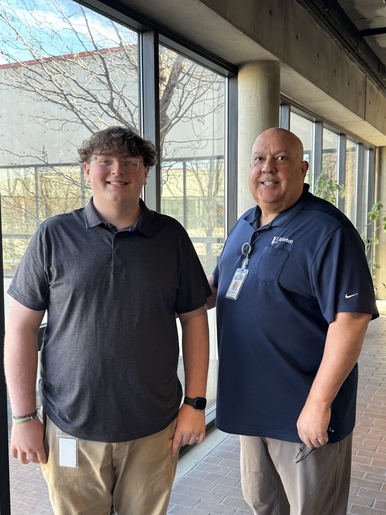Two individuals stand in a building lobby near a staircase and a sign that reads “For privacy reasons, please wait here until called,” both smiling at the camera.