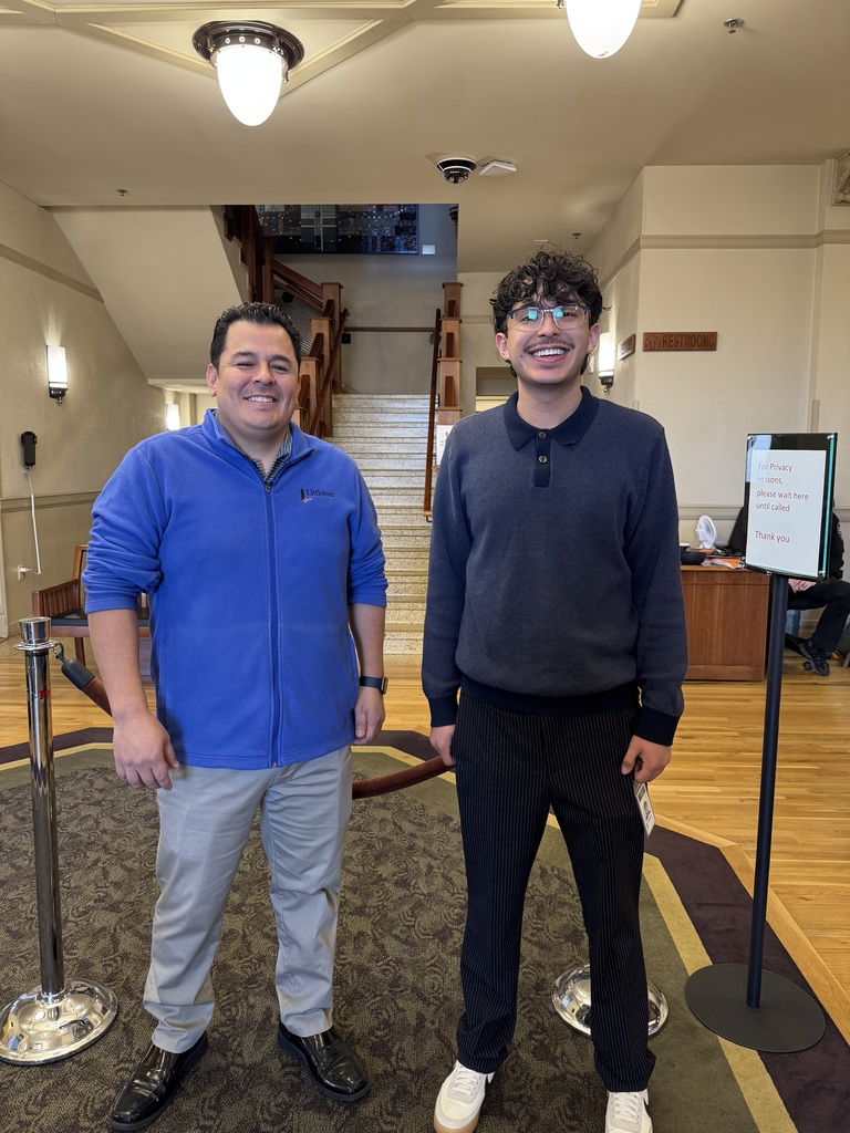 Two individuals stand in a building lobby near a staircase and a sign that reads “For privacy reasons, please wait here until called,” both smiling at the camera.