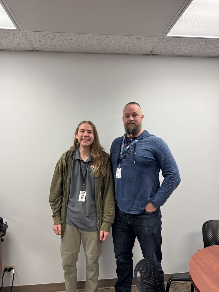 Student and adult staff member stand side by side against a plain wall, both smiling and wearing ID badges in a classroom or office setting.