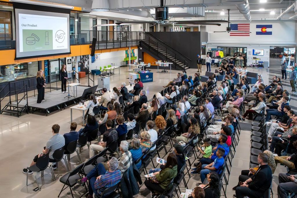 Large audience of students and adults sits in rows inside a modern school commons area, watching two students present on a stage with a slide titled “Our Product” projected on a screen.