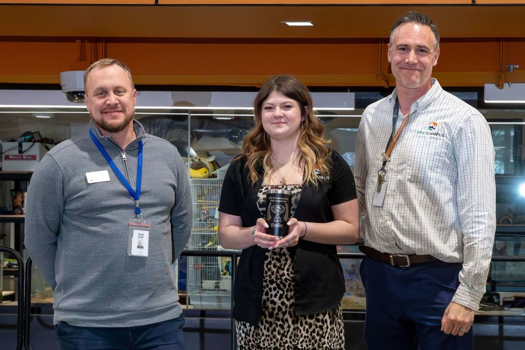 Small group of high school students gathers around an instructor, adjusting and testing clip-on microphones and audio equipment in a classroom or lab setting.
