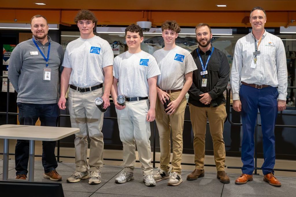Four students in matching gray shirts stand with two staff members, smiling for a group photo in a school lab or presentation space.
