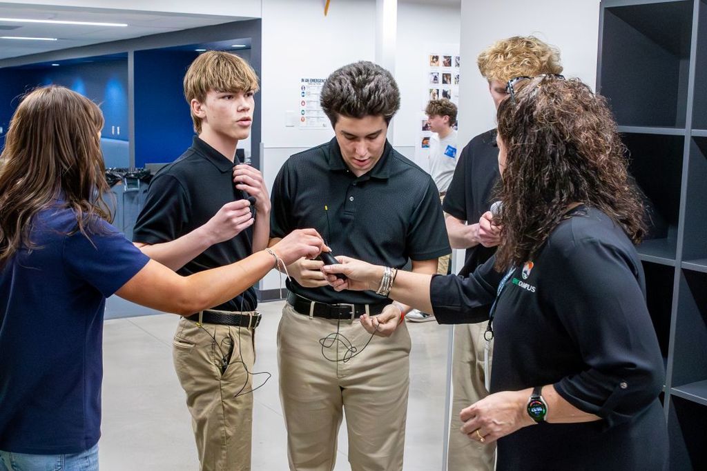 Small group of high school students gathers around an instructor, adjusting and testing clip-on microphones and audio equipment in a classroom or lab setting.
