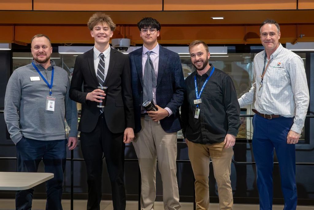 Five individuals, including two students in formal attire, stand together smiling and holding tumblers, likely after a presentation or competition.