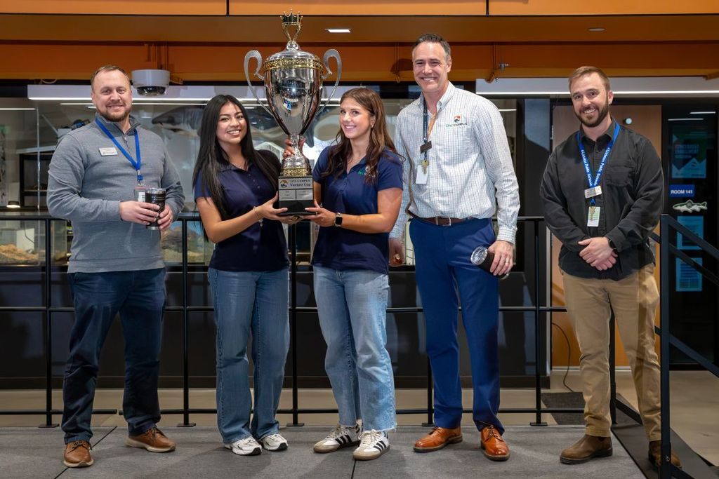 Two students hold a large trophy while standing with three staff members, smiling in a school lab space.