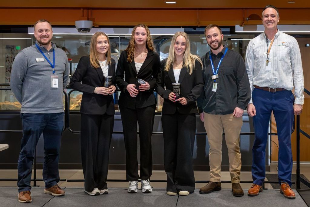 Three female students in business attire stand with two staff members, smiling and holding tumblers, posing after a presentation.