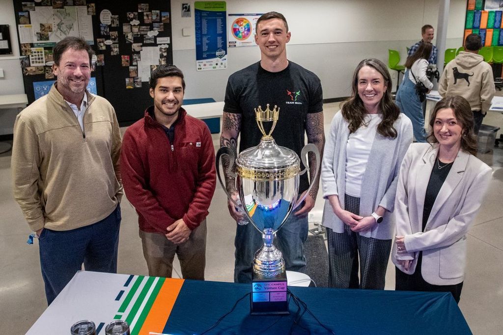 .Five people, including students and staff, stand behind a table smiling while presenting a large silver trophy labeled “EPIC Campus Venture Cup,” in a classroom or school commons space with posters and displays in the background.