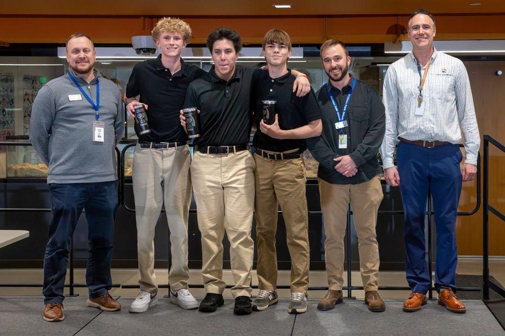 Group of students and staff stands together on a stage, smiling, with several students holding awards or tumblers.