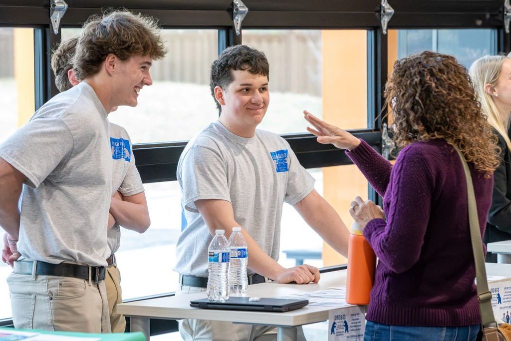 Two students stand at a table speaking with an adult, gesturing during a conversation, with water bottles and materials on the table in a classroom or event setting.