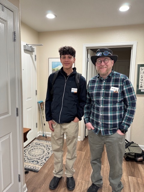 Two individuals stand side by side indoors, both smiling at the camera. One wears a black jacket and khaki pants with a backpack, while the other wears a plaid shirt, green pants, and a hat with goggles. Name tags are visible on both.