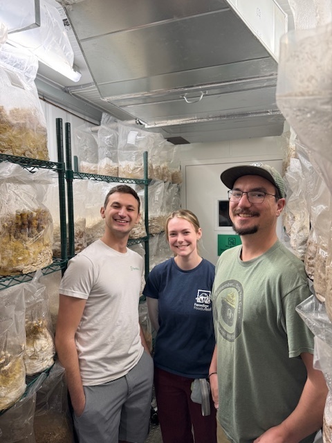 Three people stand closely together in a narrow growing room lined with shelves of bagged materials, likely mushrooms or plants. All are smiling, wearing casual work attire.