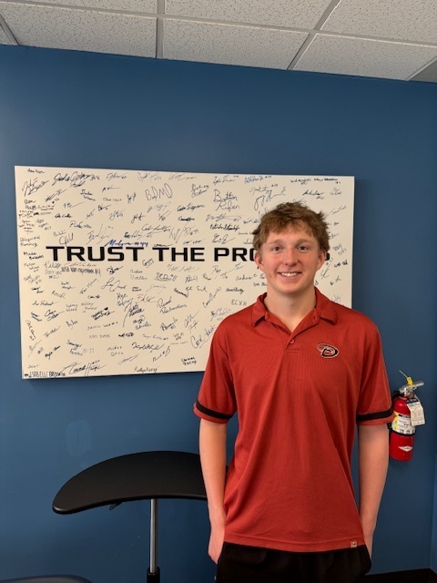 A student in a red polo shirt stands smiling in front of a wall display filled with signatures and the phrase “TRUST THE PRO…” partially visible. A small round table and a fire extinguisher are nearby.