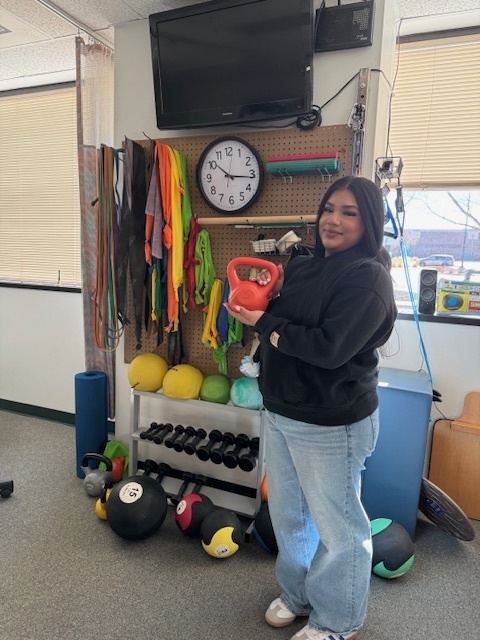 A student stands in a fitness or therapy room holding a red kettlebell. Behind them are racks of colorful exercise equipment, including resistance bands, medicine balls, and dumbbells, along with a wall clock and mounted TV.