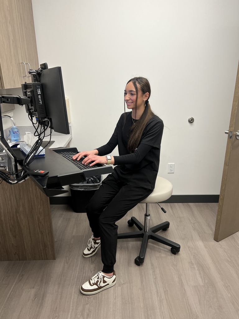 A young woman with long dark hair sits on a rolling stool in a small clinical or office room, typing on a desktop computer mounted to the wall. She wears black scrubs and sneakers, smiling as she works. A cabinet, sanitizer bottle, and keyboard tray are visible nearby.