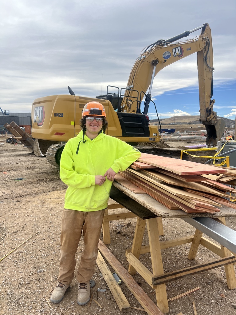 A student wearing a bright neon yellow hoodie, work pants, and an orange hard hat stands at a construction site, smiling and leaning on a wooden worktable. A large CAT excavator and construction materials are visible behind them under a cloudy sky.