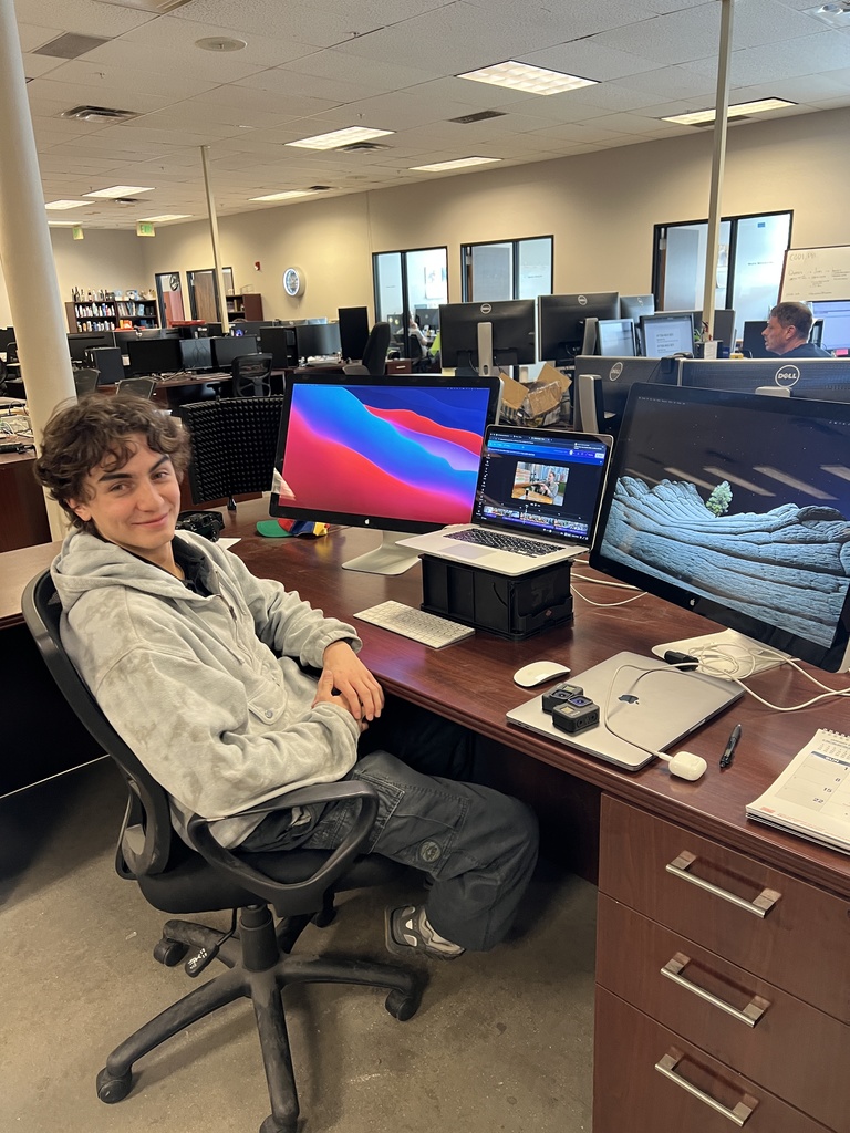 A student with short curly hair sits at a desk in an open office workspace, smiling at the camera. Multiple computer monitors, a laptop, and editing software are visible on the desk, with rows of workstations and colleagues in the background.