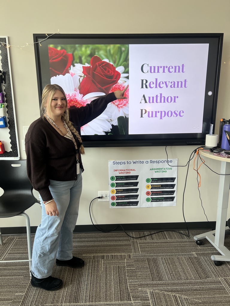 A female teacher stands in a classroom pointing at a large screen displaying the words “Current Relevant Author Purpose” beside an image of flowers. A poster below the screen outlines steps to write a response.