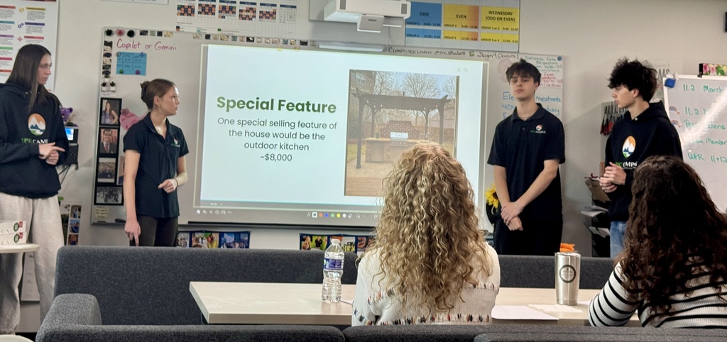 Students stand in front of a classroom presentation slide titled “Special Feature,” which reads: “One special selling feature of the house would be the outdoor kitchen ~$8,000.” An image of an outdoor kitchen is displayed, while audience members sit at a table in the foreground.