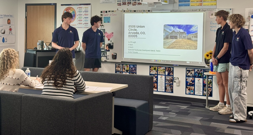 Four high school students in matching navy polos stand at the front of a classroom presenting to two seated adults. A projected slide shows a house listing with the address “8506 Union Circle, Arvada, CO, 80005,” along with details like square footage, bedrooms, bathrooms, and features including a backyard deck and Tesla solar panels.