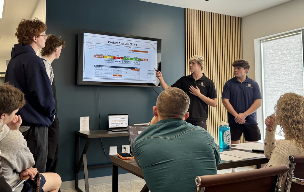 In the same meeting space, a student points to a screen showing a “Project Analysis Sheet” with financial figures and charts. Other students stand nearby while seated adults review the presentation, with laptops, notebooks, and a water bottle on the table.