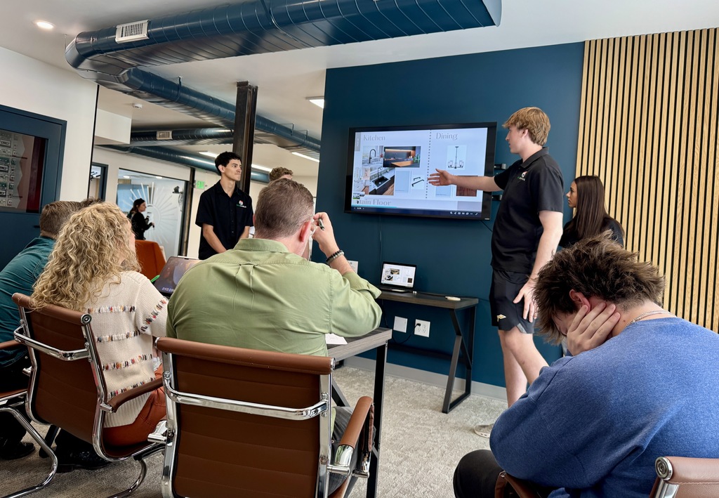 Students present in a modern meeting room with teal walls and wood paneling. One student gestures toward a large screen displaying a home design layout labeled “Kitchen” and “Dining,” while several adults sit in chairs watching and taking notes.