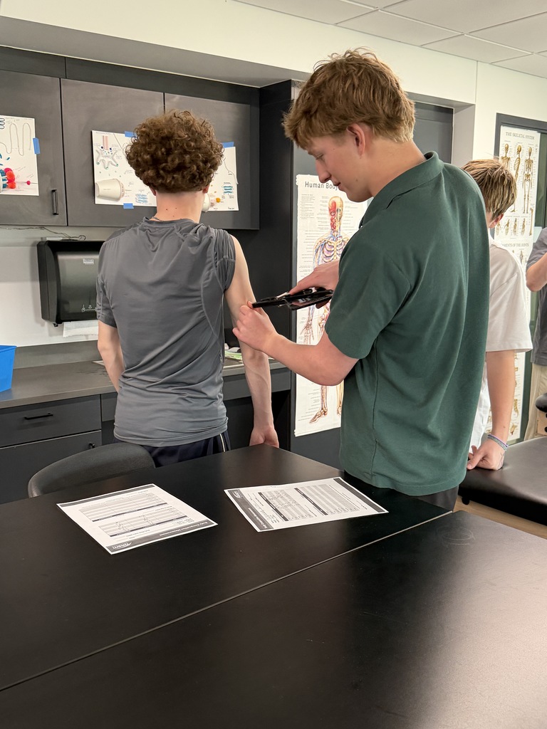 A student measures another student’s arm using calipers while a third student observes. Papers with recorded data lie on the table, and anatomical posters and lab equipment are visible in the classroom, suggesting a hands-on health or sports science activity.