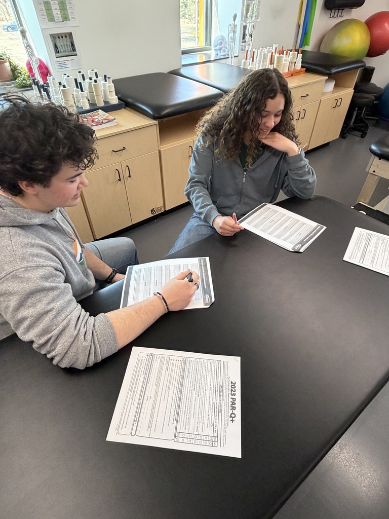 Two students sit at a table in a health sciences classroom, reviewing and filling out forms labeled “2023 PAR-Q+.” Therapy tables, anatomical models, and exercise equipment are visible in the background, indicating a fitness or rehabilitation learning environment.