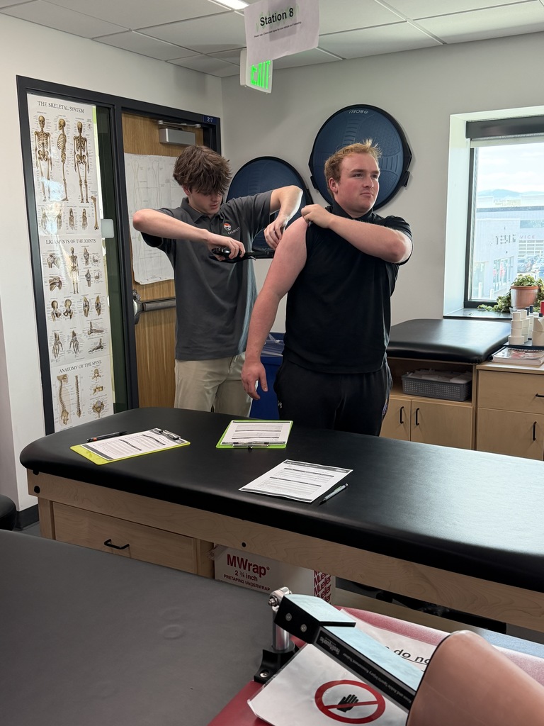 One student uses skinfold calipers to measure another student’s upper arm in a classroom lab setting. A sign reading “Station 8” hangs above, and anatomy posters and clipboards with worksheets are visible around the room.