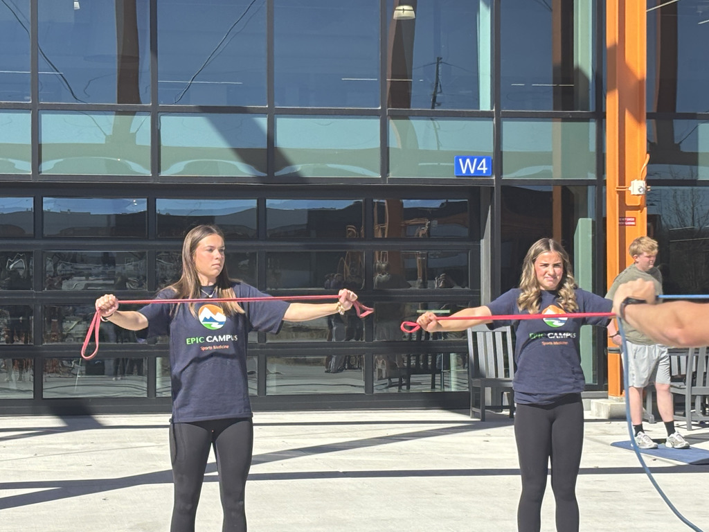 Two teenage girls stand outdoors in front of a glass school building labeled “W4,” each holding a red resistance band stretched across their chest. Both wear navy “EPIC Campus Sports Medicine” T-shirts and black leggings, focusing forward as they exercise.