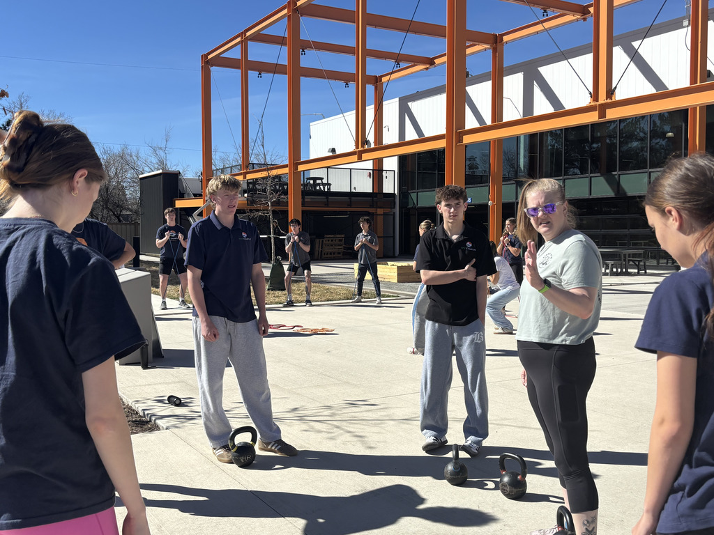 A group of diverse high school students stands in a circle on a concrete courtyard under an orange steel canopy. A female student in a light green shirt appears to be speaking or instructing while others listen; kettlebells and resistance bands are placed on the ground.