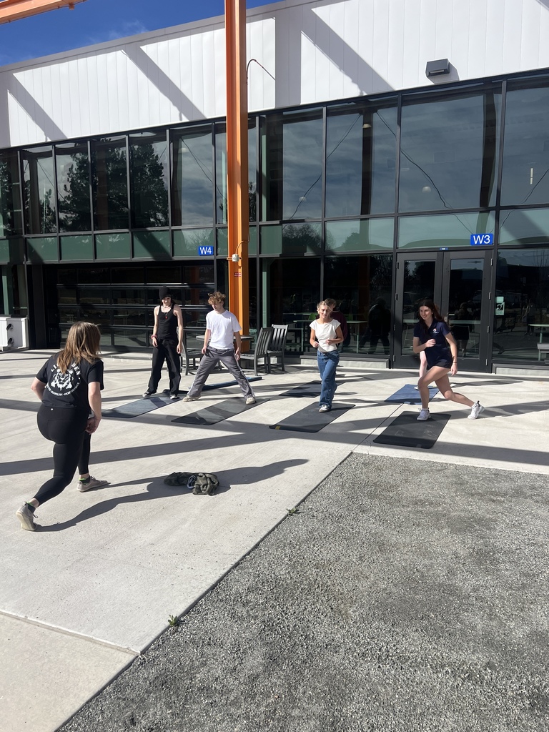 Several students exercise on black mats outside the school near doors labeled “W3” and “W4.” They appear to be doing lunges or balance exercises while a classmate or instructor stands nearby observing.
