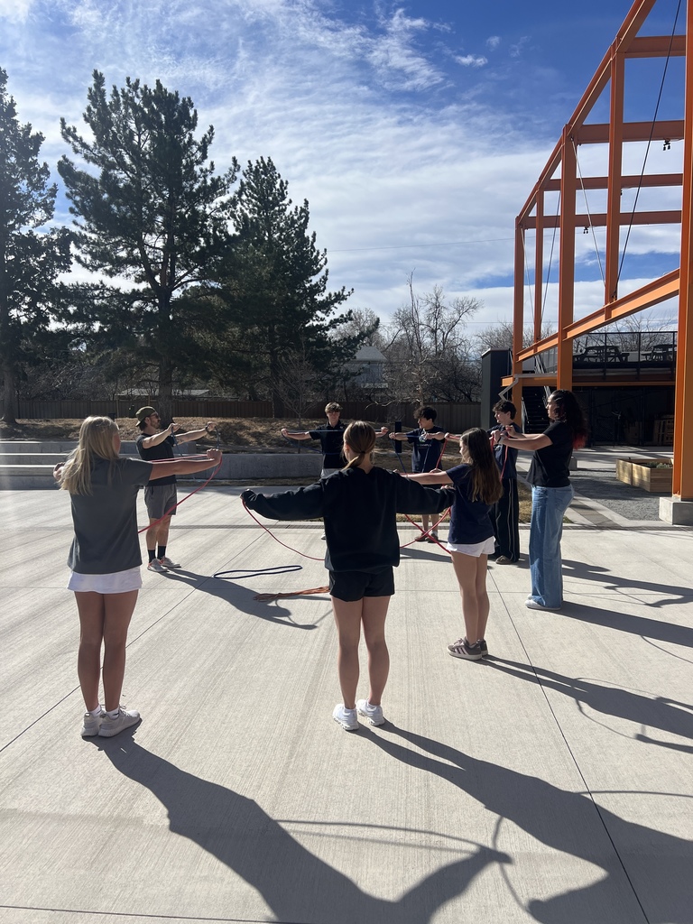 Students form a wide circle, each holding resistance bands extended outward while facing inward. They stand on a concrete courtyard surrounded by trees and the orange canopy structure.