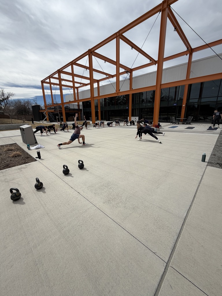 A large group of students participates in a synchronized stretching or lunge exercise across the courtyard. Multiple kettlebells are lined up in the foreground, and the orange steel canopy spans overhead.