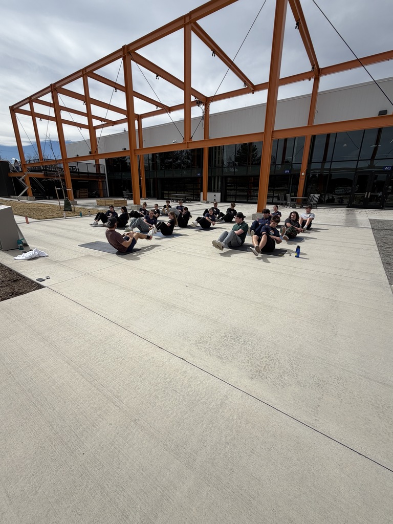 A class of students sits on the ground in rows performing a core exercise, leaning back with legs raised. The outdoor courtyard and orange canopy structure extend behind them under a lightly overcast sky.