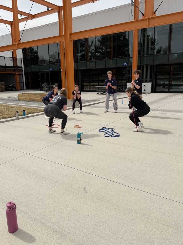 A small group of students performs squat exercises with resistance bands. Water bottles and bands are placed on the ground around them, and the school building with large windows is in the background.