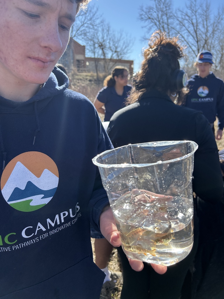 A teenage student in a navy EPIC Campus hoodie looks down at a clear container of water with several small fish inside. Other students stand in the background outdoors near a natural area with trees and dry grass.