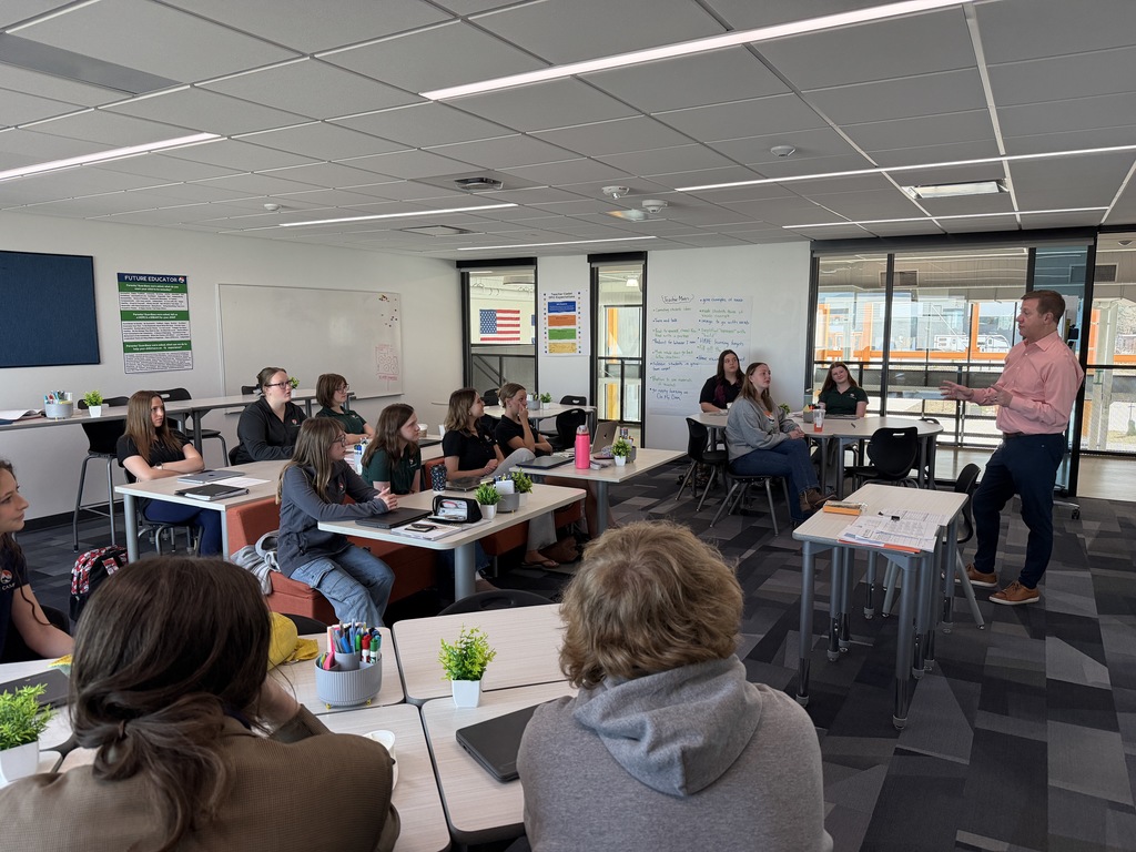 A classroom scene with a male teacher in a pink button-down shirt speaking at the front while gesturing with his hands. Students of diverse backgrounds sit at grouped tables, listening attentively with laptops, notebooks, and water bottles visible. A whiteboard behind the teacher displays handwritten notes, and posters—including one labeled “Future Educator”—hang on the walls.