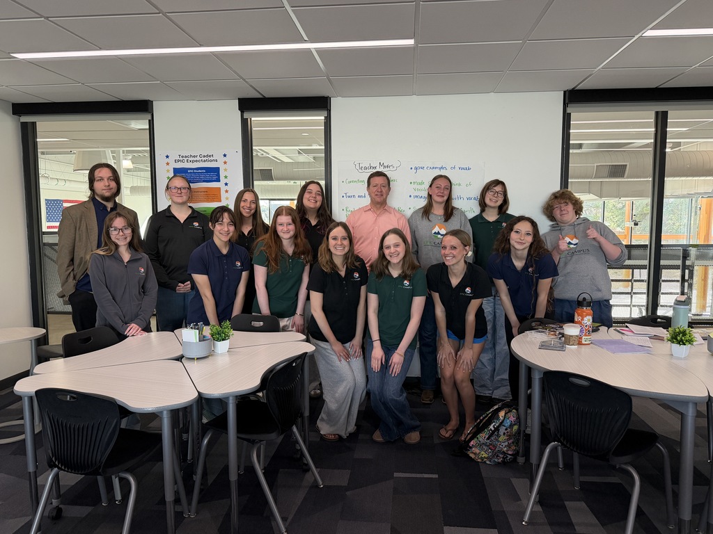A group of about 14 high school students and one adult stand together in a classroom, smiling for a group photo. Students wear EPIC Campus polos and casual attire. Behind them are whiteboards with handwritten notes, a poster titled “Teacher Cadet EPIC Expectations,” and large interior windows. Tables with small plants, notebooks, and water bottles are arranged in the foreground.