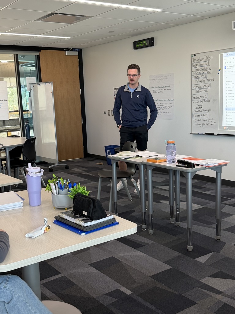 Teacher stands at the front of a modern classroom beside a desk, speaking to students. He appears to be a young adult male with light skin, short brown hair, glasses, and a trimmed mustache, wearing a navy pullover and black shorts. His hands are in his pockets as he faces the class. On the desk in front of him are papers, a reusable water bottle, a stapler, and folders.