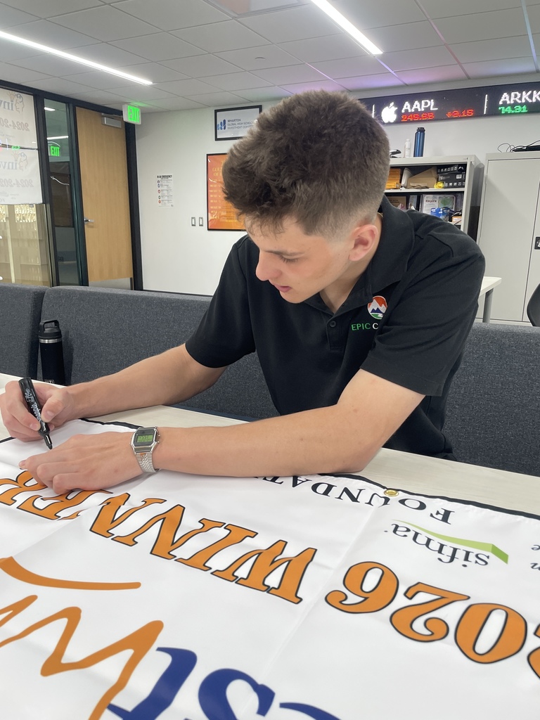 A teenage male student in a black EPIC Campus polo sits at a table, signing a white “investWrite 2025–2026 WINNER” banner with a black marker. He leans forward, focused, wearing a silver watch. In the background are a classroom/office space, shelves, and an electronic stock ticker display showing symbols like “AAPL.”
