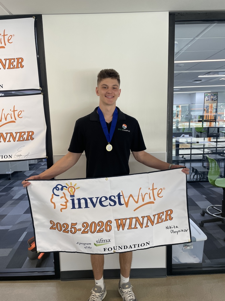 A teenage male student wearing a black EPIC Campus polo and a medal on a blue ribbon stands holding a white banner that reads “investWrite 2025–2026 WINNER,” with a handwritten name visible on the right side. He smiles at the camera in a school office setting with glass walls, chairs, and classroom furniture behind him.