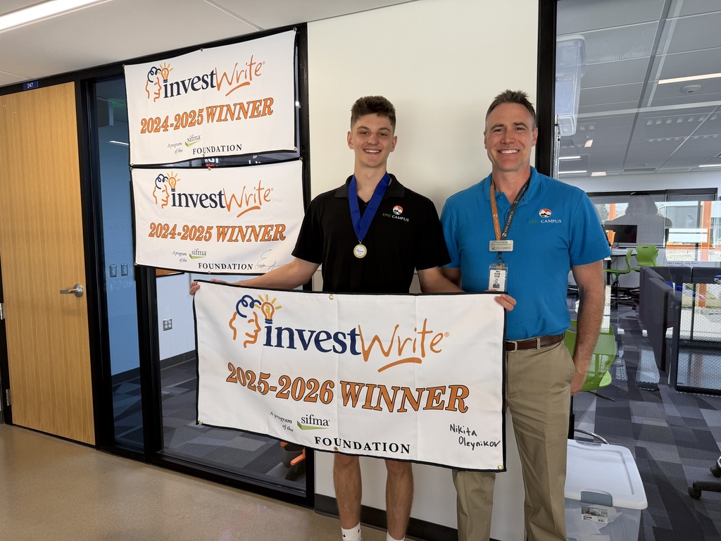 Two people—a teenage male student and an adult male staff member—stand smiling in a modern school office space, holding a white banner that reads “investWrite 2025–2026 WINNER.” The student, wearing a black polo with an EPIC Campus logo and a medal on a blue ribbon, stands beside the staff member in a teal polo and ID badge. Behind them, two additional banners read “investWrite 2024–2025 WINNER.” Glass walls, office furniture, and a classroom setting are visible in the background.